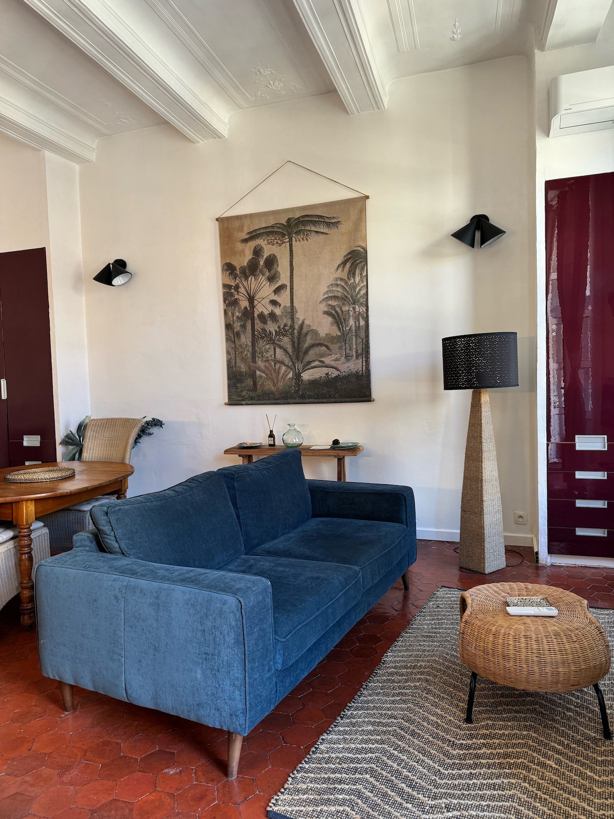 A comfortable living area features a plush blue sofa with wooden legs, positioned across from a wooden dining table. A woven rug adds texture to the space. A decorative wall hanging and a standing lamp provide visual interest, along with a small side table displaying decorative items.