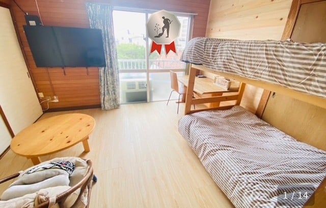 A cozy room features a bunk bed with a checkered comforter and a spacious wooden table. Natural light fills the space through a sliding door that opens to a balcony. A television is mounted on the wall opposite the bed, enhancing the functional layout.