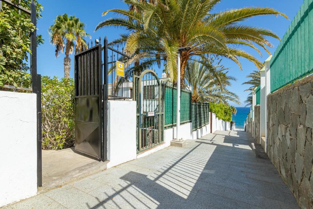 A pathway is seen leading to the beach, flanked by palm trees and greenery. A black metal gate is positioned at the entrance, with sunlight casting shadows on the stone pavement. The ocean is visible at the end of the walkway, suggesting proximity to the shoreline.