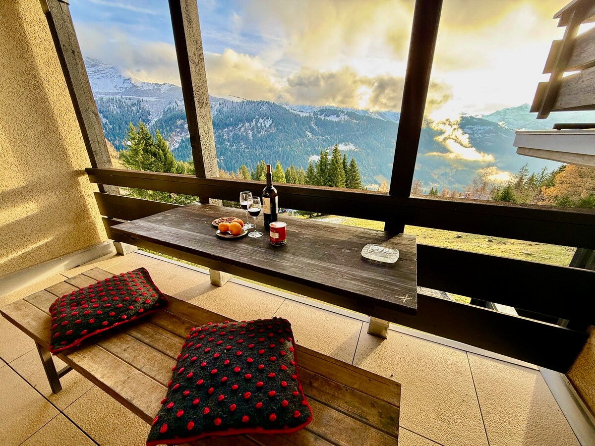 A wooden table is set on a balcony, featuring a bottle of wine, glasses, and a plate with fruit. Two cushions with red polka dots are arranged on the bench. The breathtaking view of mountains and valleys is visible in the background, complemented by a colorful sky.