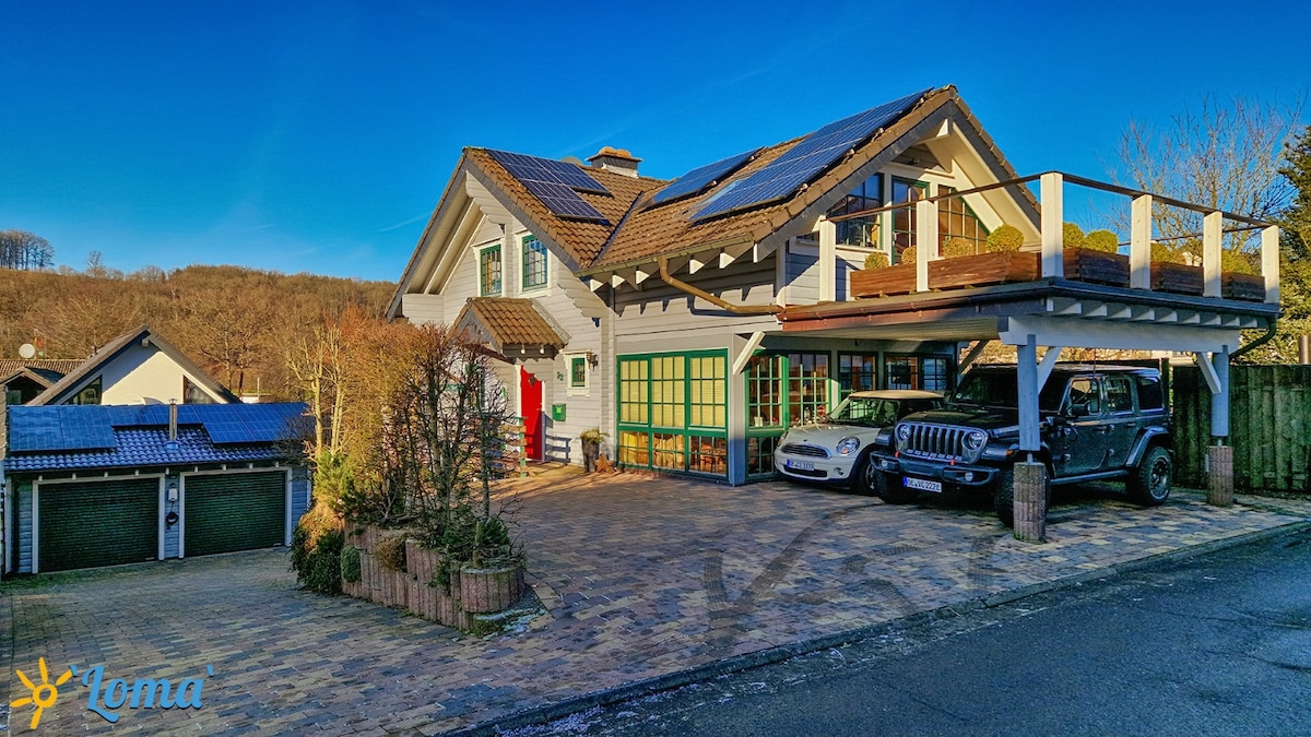 The exterior of the property showcases a multi-story house with solar panels on the roof. A spacious driveway is visible, featuring two parked vehicles. Surrounding greenery adds to the serene environment, with clear blue skies enhancing the overall appearance.