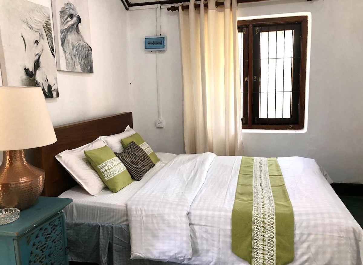 A serene sleeping area features a spacious bed dressed in white linens adorned with a green decorative runner. Natural light filters through a small window with a grid pattern, and two textured artworks are displayed on the wall, adding a touch of elegance.