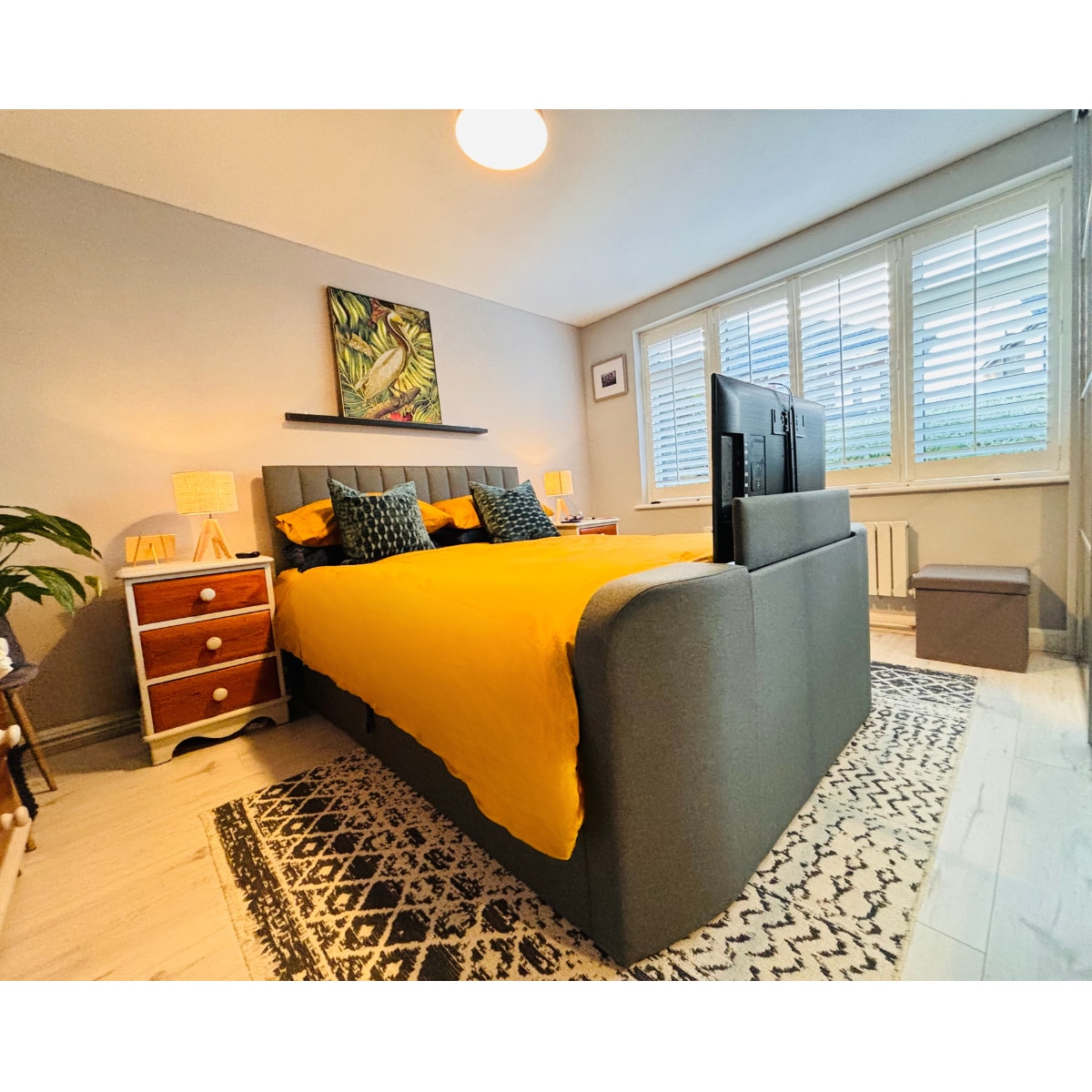 The bedroom features a spacious bed dressed in vibrant yellow bedding. Two bedside tables with lamps are positioned on either side. A large piece of art adorns the wall above the bed, and natural light is filtered through shuttered windows. A cozy rug lies beneath.