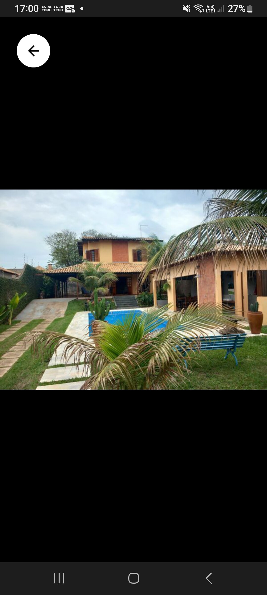 A spacious backyard is surrounded by well-maintained greenery, featuring a stone path leading to the home. Two palm trees offer tropical charm, while a blue seating area invites relaxation under the sun. The two-story house showcases large windows and a terracotta-styled roof.
