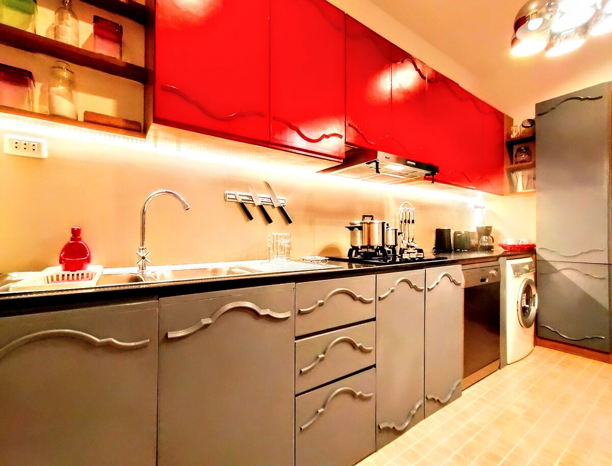 A modern kitchen is displayed, featuring sleek grey cabinetry with artistic wave designs and vibrant red upper cabinets. The area includes a stainless steel sink, built-in stove, and a washing machine. Ample countertop space is complemented by warm under-cabinet lighting.