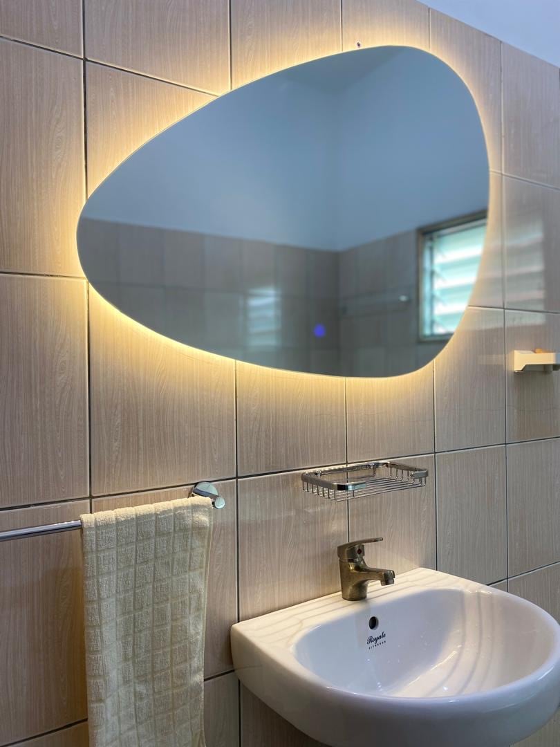 A modern bathroom features a uniquely shaped illuminated mirror above a sink. Soft lighting accentuates the mirror's edges, while the wall is tiled in a neutral shade. A towel hangs neatly nearby, and a small shelf holds toiletries, providing a functional and stylish space.