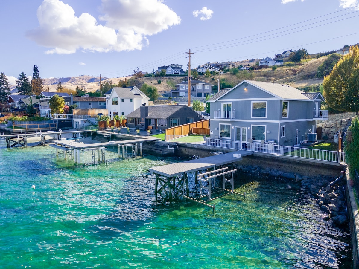 This waterfront home is surrounded by clear turquoise waters, featuring docks for boats along the shoreline. The two-story structure showcases large windows, inviting natural light and offering stunning views of the lake and nearby hills.