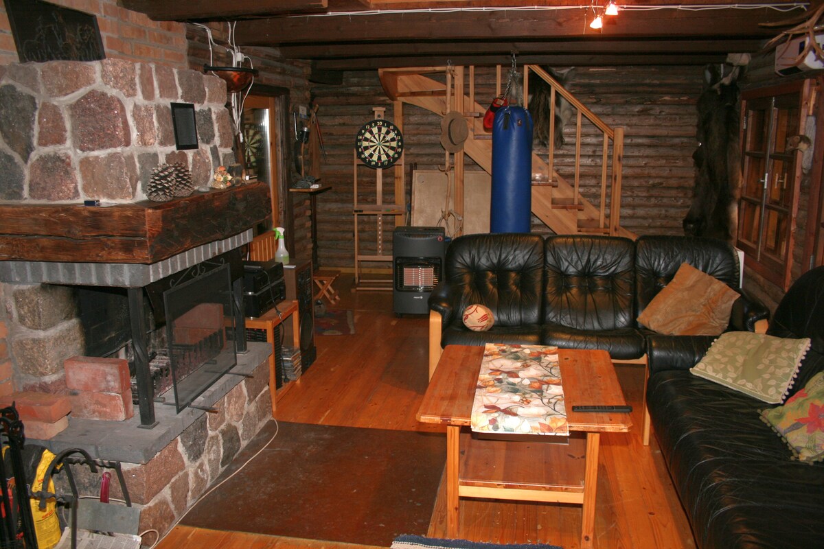 A cozy living area features a stone fireplace as the focal point, surrounded by comfortable seating. A wooden coffee table occupies the center, adorned with a decorative table runner. A staircase leads to the upper level, while a dartboard and additional amenities provide options for entertainment.