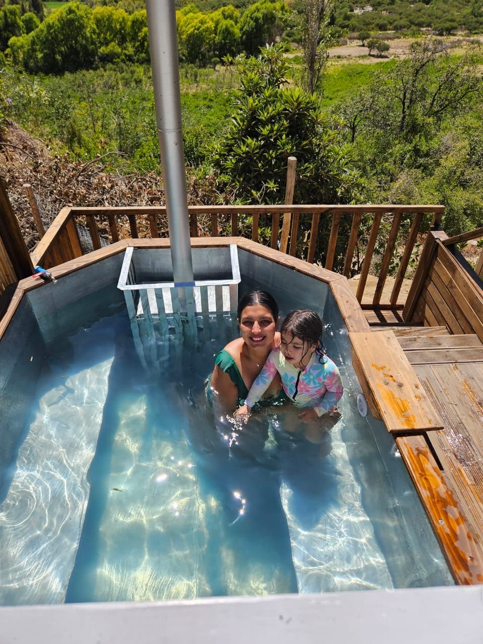 A wooden hot tub is situated on a deck, filled with clear water that reflects sunlight. Lush greenery surrounds the area, providing a serene backdrop. Steps lead down from the tub, indicating access to the outdoor space.