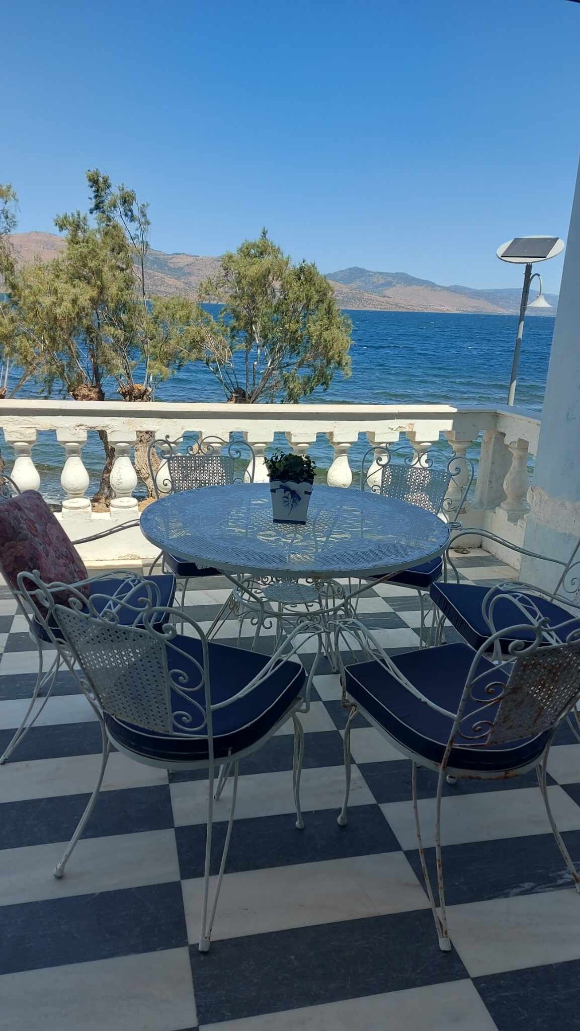 An outdoor dining area is seen with a round table surrounded by four chairs, featuring a classic design. The setting offers views of the water and distant mountains, framed by lush trees. A calm blue sky complements the serene atmosphere.