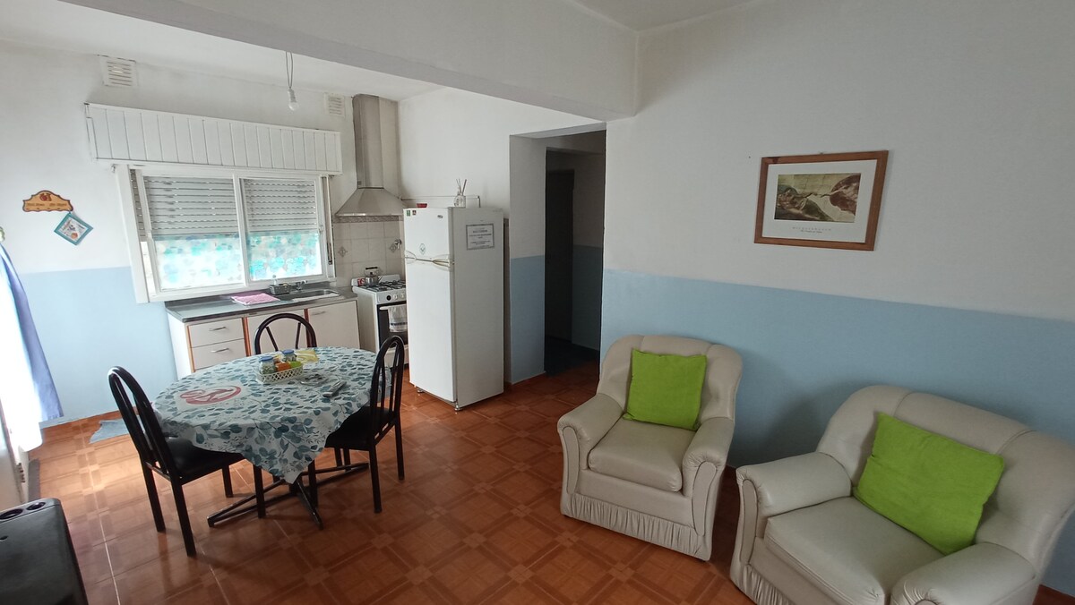 A welcoming dining area features a round table adorned with a floral tablecloth, surrounded by four chairs. Two comfortable armchairs with green cushions are positioned near a window, providing natural light. A kitchenette is visible in the background, alongside a refrigerator and a small oven.