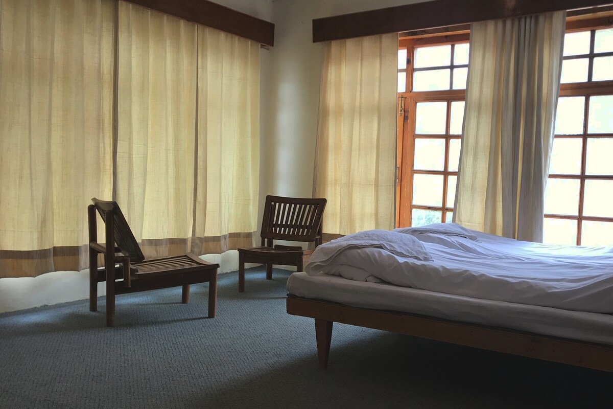 A spacious sleeping area features a large bed with a neatly arranged white duvet. Two wooden chairs are positioned near bright, large windows, allowing natural light to filter through light-colored curtains.