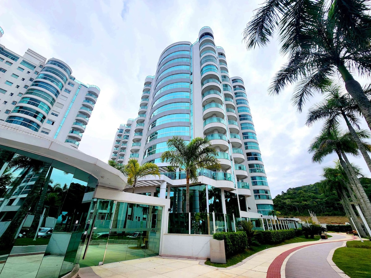 The exterior of a modern high-rise building is visible, showcasing large glass windows and curved balconies. Lush palm trees and well-maintained landscaping frame the entrance, leading to a welcoming pathway. The surrounding hills add depth to the scenic view.