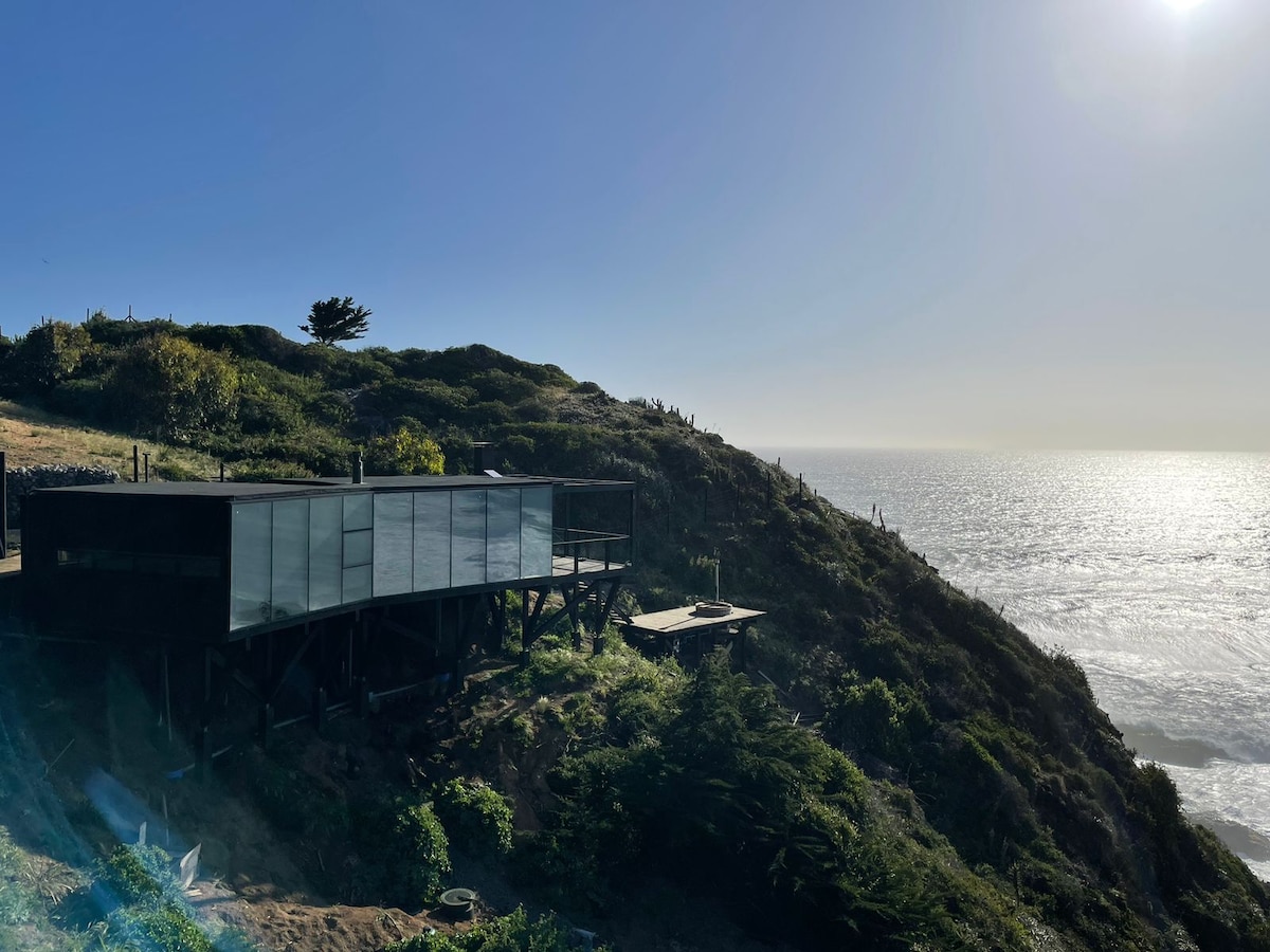 A modern cabin is nestled on a hillside, with large glass windows reflecting the sunlight. The structure offers expansive views of the ocean and surrounding greenery. The terrain slopes down toward the water, providing a natural, serene setting.