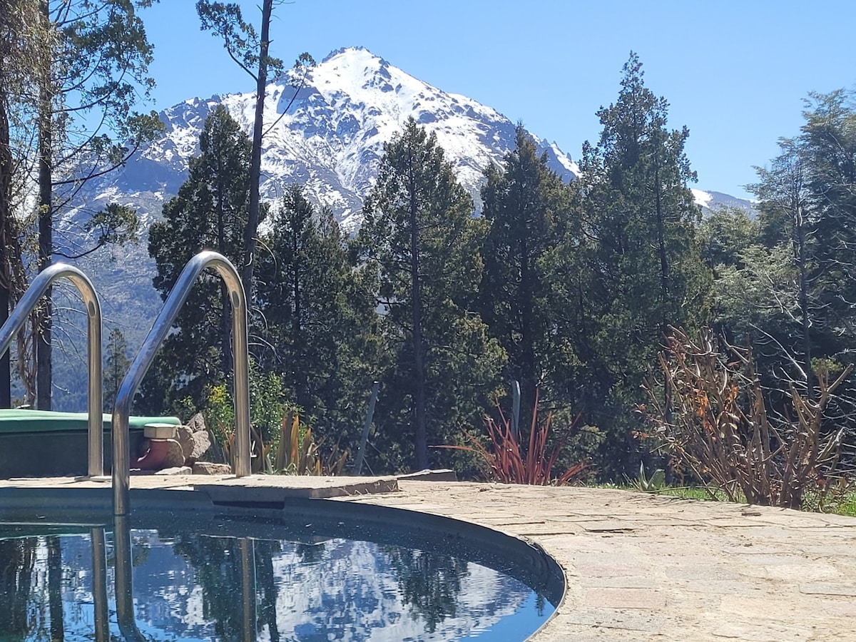 A serene pool reflects the surrounding landscape, including tall trees and a snow-capped mountain in the background. Clear blue skies enhance the tranquil atmosphere, emphasizing the natural beauty of the environment.