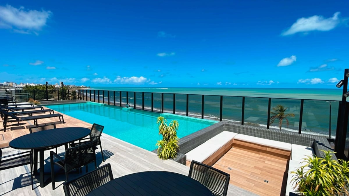 An inviting rooftop area features a clear swimming pool with a wet deck, framed by sleek glass railings. Sun loungers are arranged along the deck, offering a serene view of the ocean and sky. Lush greenery adds a touch of nature to the modern design.