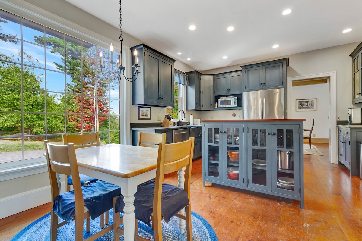 A bright kitchen features dark cabinetry and stainless steel appliances. A wooden dining table with four light-colored chairs is positioned nearby, complemented by a blue area rug. Large windows invite natural light, revealing views of the surrounding outdoor greenery.