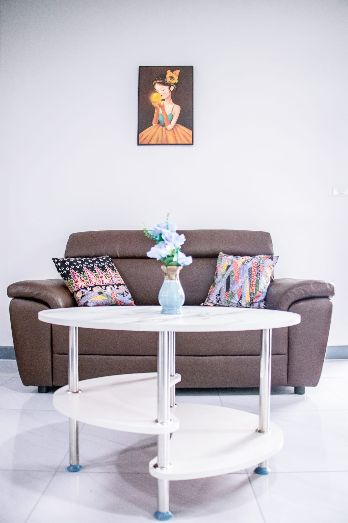 A cozy seating area is highlighted by a brown sofa adorned with decorative pillows. A circular coffee table, featuring three tiers, is positioned centrally, topped with a vase holding blue flowers. A colorful artwork is displayed on the light gray wall above the sofa.