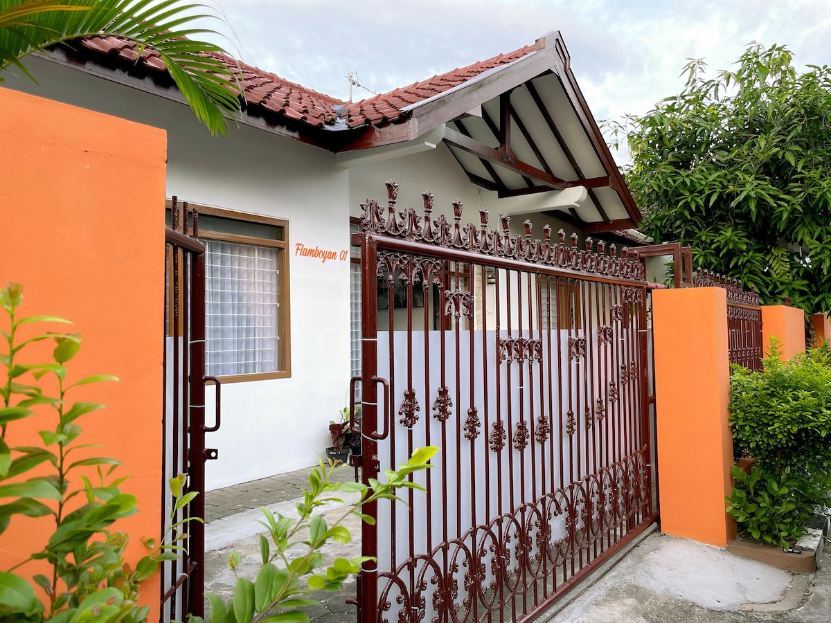A welcoming entrance is framed by a sturdy, decorative iron gate and vibrant orange walls. Lush greenery surrounds the pathway leading to the house, while a window with a delicate curtain offers a glimpse into the interior. The roofline features traditional architecture.