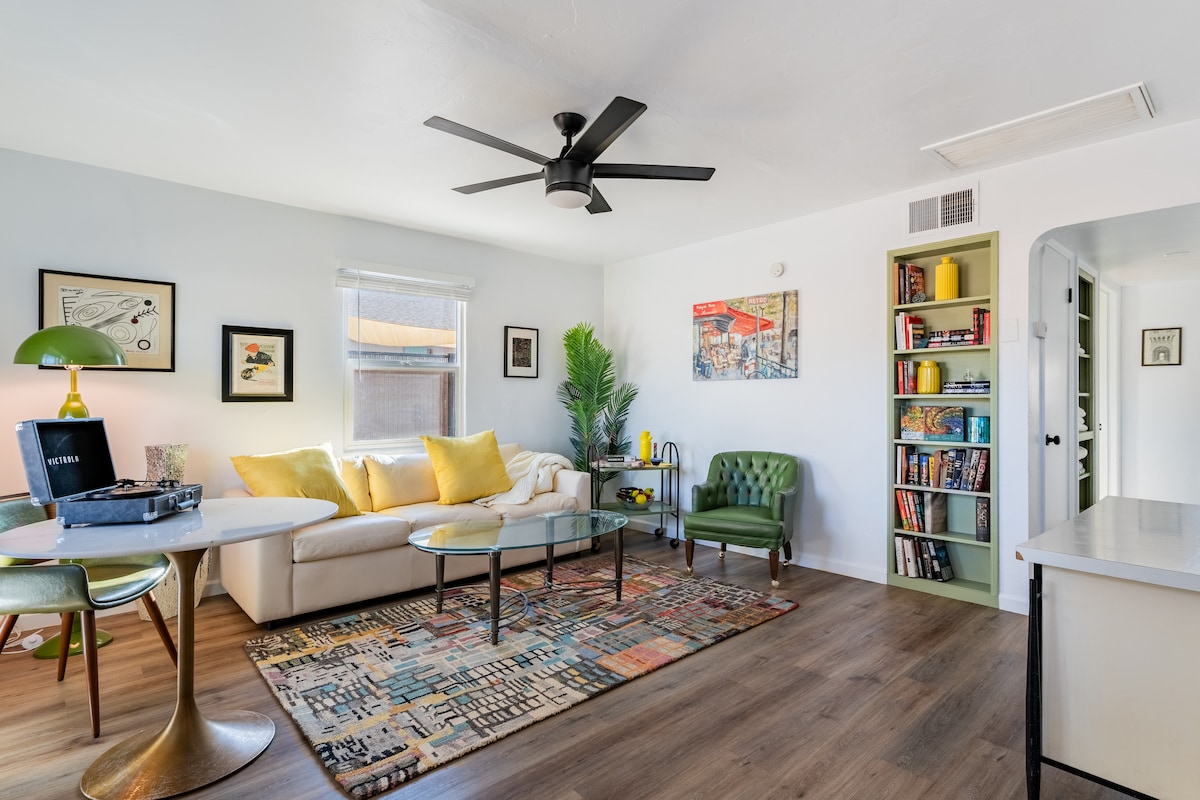 A bright living area features a comfortable white sofa adorned with yellow cushions, accompanied by a round dining table with modern chairs. A ceiling fan provides airflow, and a bookshelf filled with colorful books adds a touch of personality. Natural light streams through a window, enhancing the inviting atmosphere.
