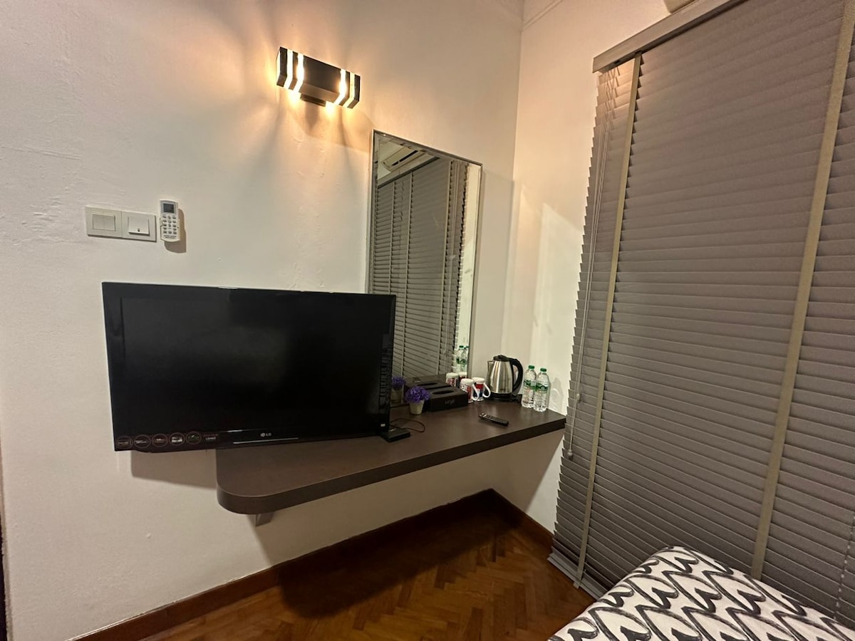 A sleek wall-mounted television sits above a minimalist ledge, which displays a kettle and two bottles of water. A large mirror reflects soft lighting from an adjacent wall sconce, creating a well-organized ambiance. Vertical blinds cover the window, allowing controlled light filtering.