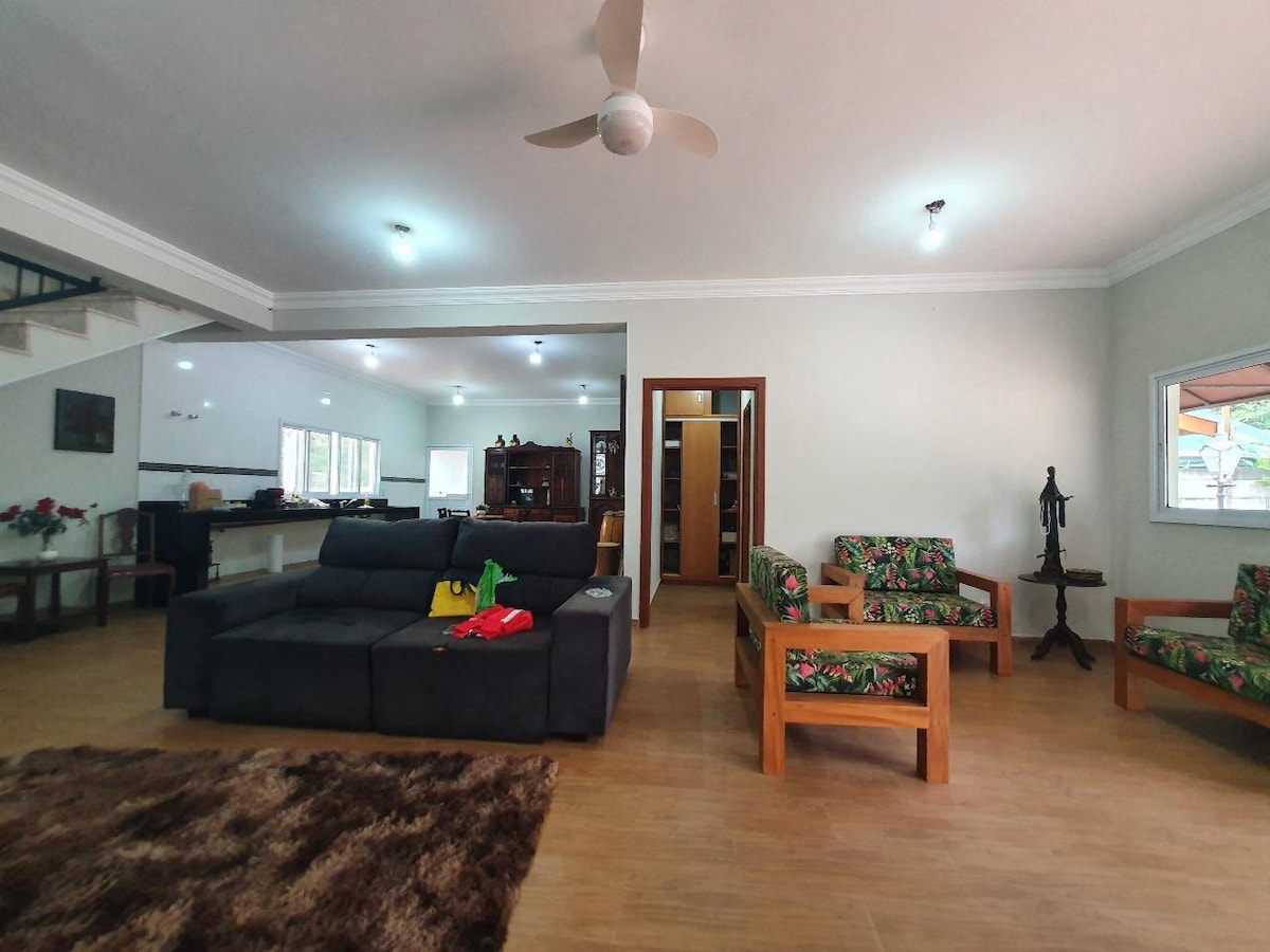 A spacious living area features a dark couch and wooden chairs adorned with bright floral cushions. Natural light enters through large windows, enhancing the openness of the space. A plush rug covers the floor, providing comfort underfoot.