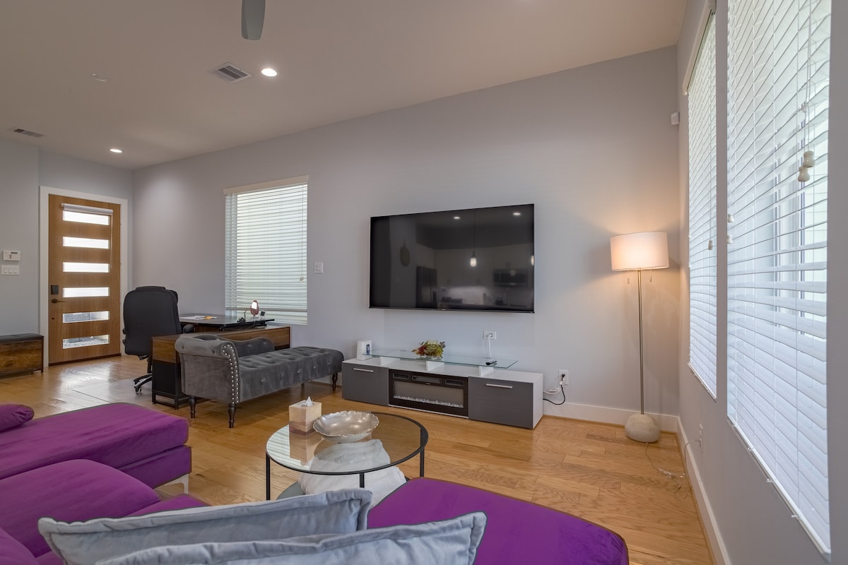 A modern living area features a comfortable purple sectional sofa arranged around a glass coffee table. A sleek entertainment unit displays a flat-screen TV, while a decorative lamp provides ambient lighting. Windows with blinds allow natural light, and a desk area is positioned in the background.