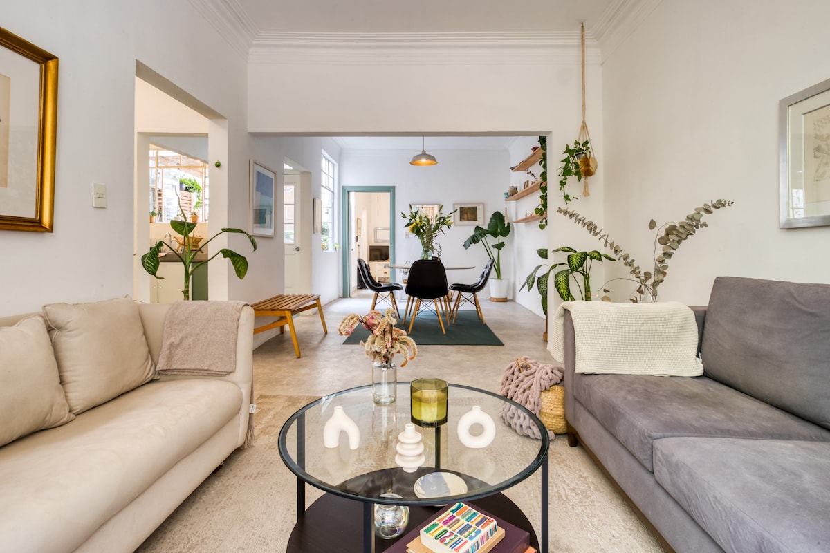 The living area features a blend of light-colored sofas, positioned around a round coffee table with decorative objects. Green plants are integrated into the space, with a view into an adjoining room showcasing dining chairs and additional greenery.