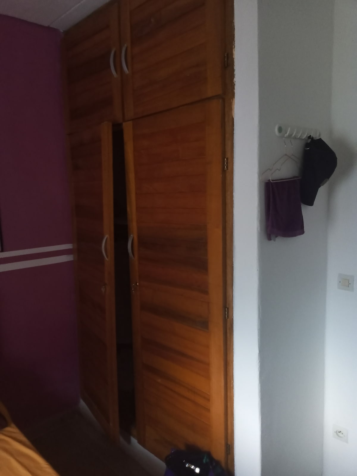 A wooden wardrobe with double doors stands against a purple wall. A small wall hook displays a hat and a piece of clothing, while a light source casts a soft glow on the wardrobe's surface, highlighting its natural finish.