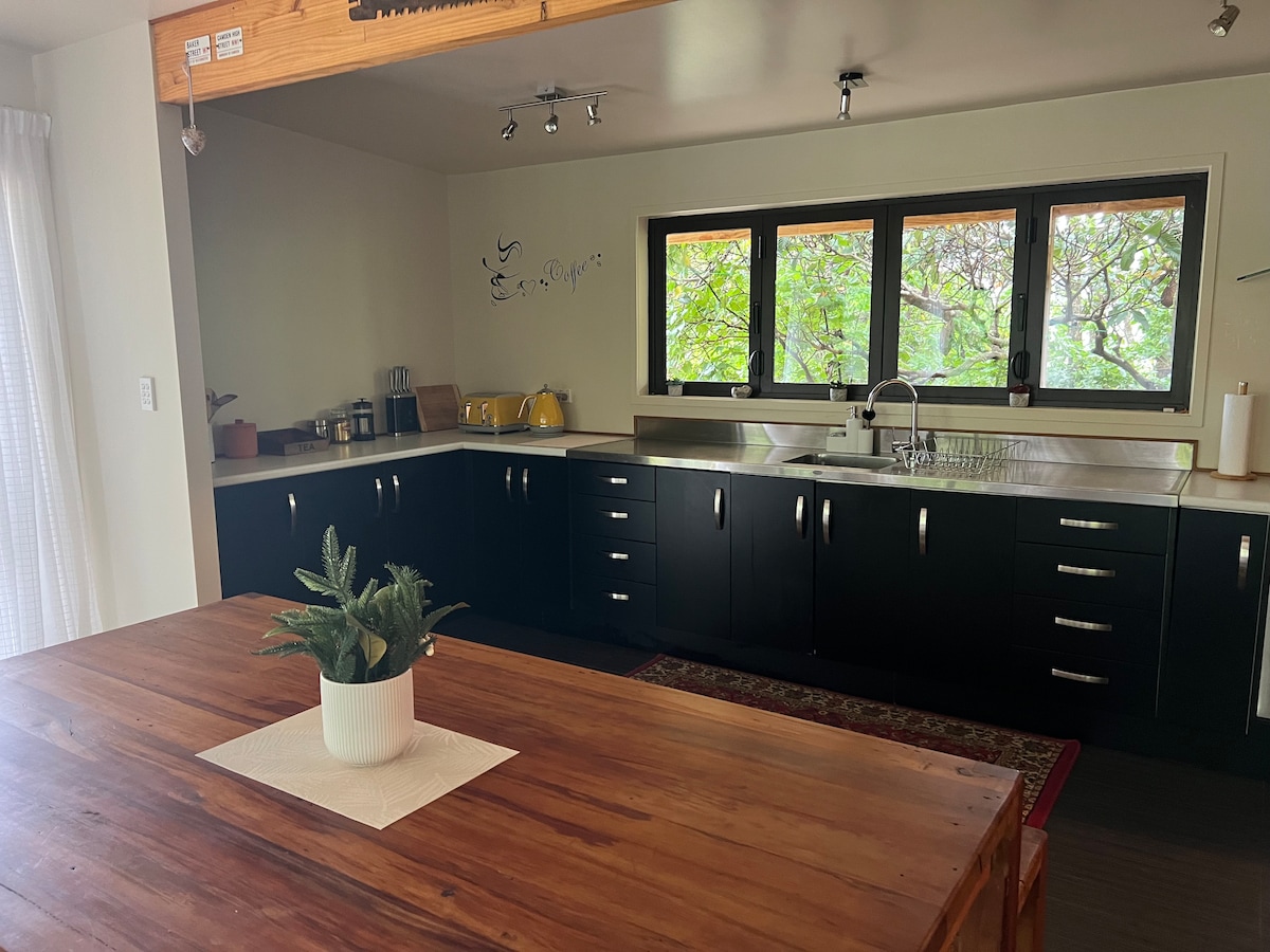 The open-plan kitchen features dark cabinetry and modern appliances, including a sink and stove. Large windows allow natural light to illuminate the space, framed by greenery outside. A wooden dining table sits at the center, complemented by a small decorative plant.
