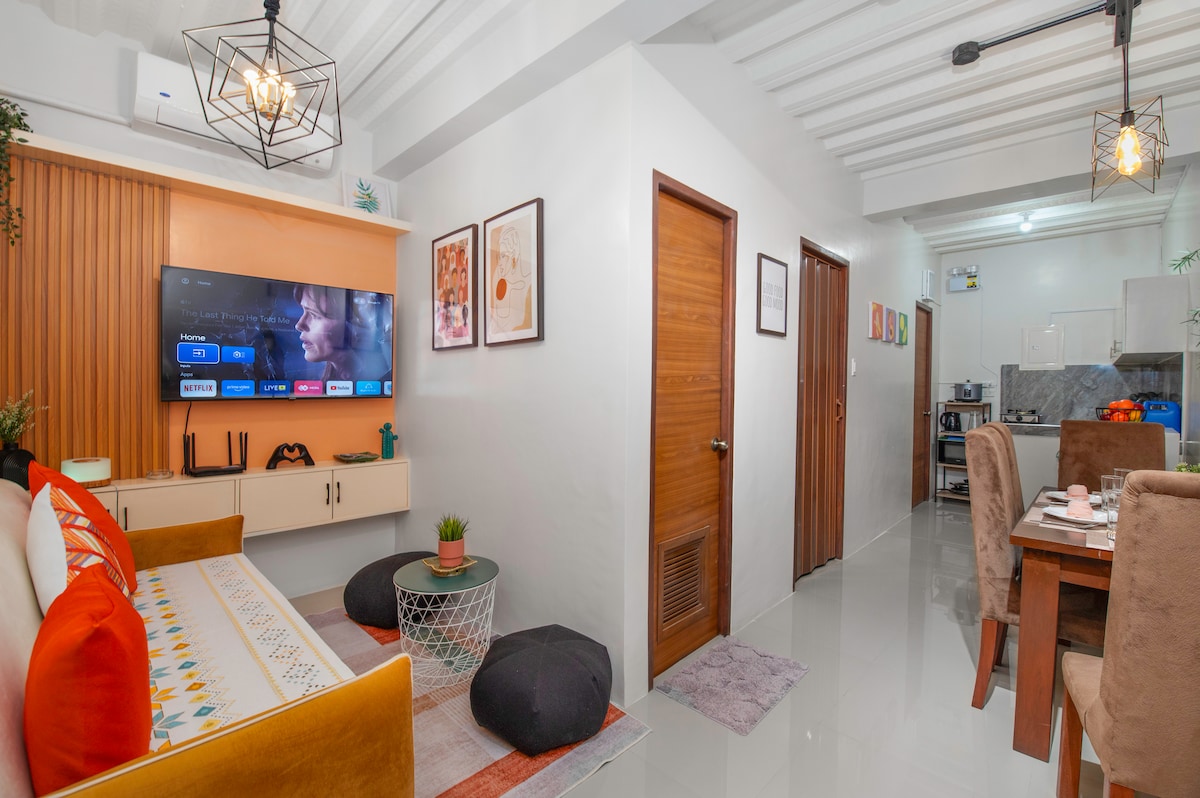 A modern living area features a long sofa with colorful cushions, a 52-inch Smart TV mounted on a wooden panel, and a small round table with potted greenery. The space is well-lit with pendant lighting and flows into a dining area that is visible in the background.