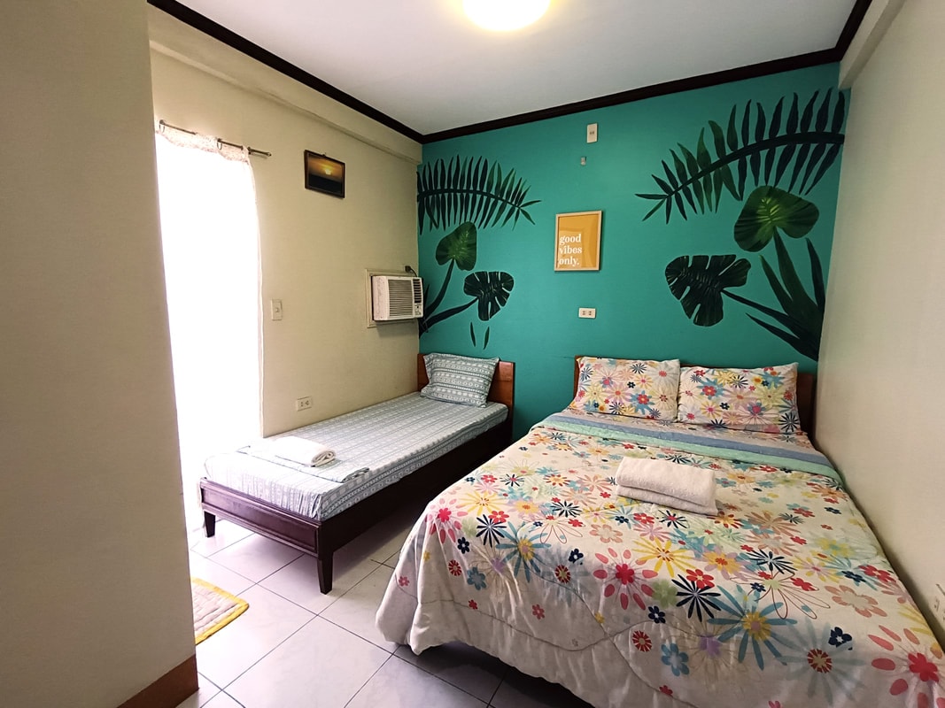 A room designed for comfort features a spacious double bed covered in a floral print blanket next to a single bed with a light blue mattress. Green leaf motifs adorn the turquoise walls, and a window allows natural light to enter the space, complemented by an air conditioning unit.