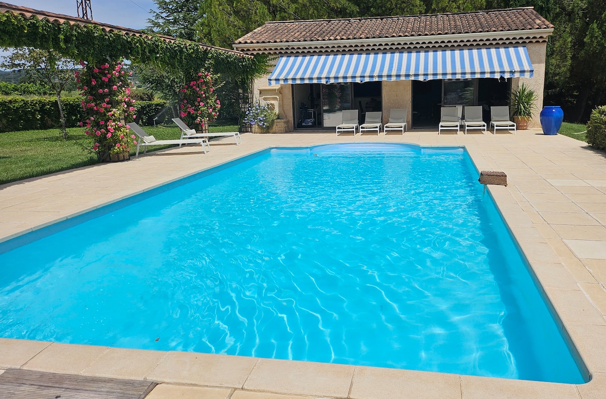 A refreshing swimming pool is featured, surrounded by a spacious tiled area. Sun loungers are positioned along one side, while a charming pool house with blue-striped awnings adds to the setting. Lush greenery and blooming flowers enhance the tranquil outdoor environment.