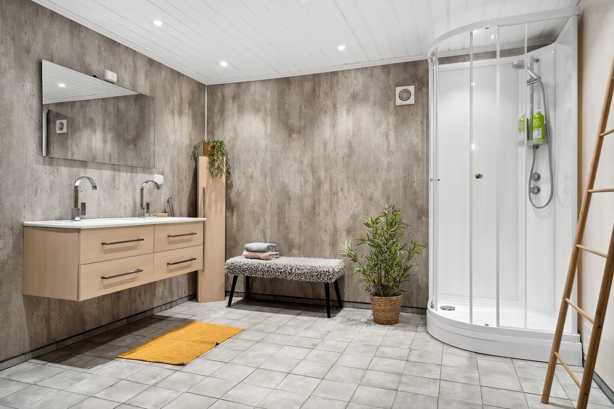 A modern bathroom features a spacious shower enclosed in glass. The neutral-toned walls complement the wooden vanity with dual sinks. A cozy bench and a small indoor plant add subtle accents, while a soft mat lies on the tiled floor.