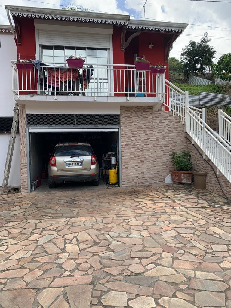 A two-story building with a red exterior features a covered balcony adorned with potted plants. A vehicle is parked in the garage on the lower level, accessible via a stone driveway. Stairs lead from the driveway to the balcony entrance.