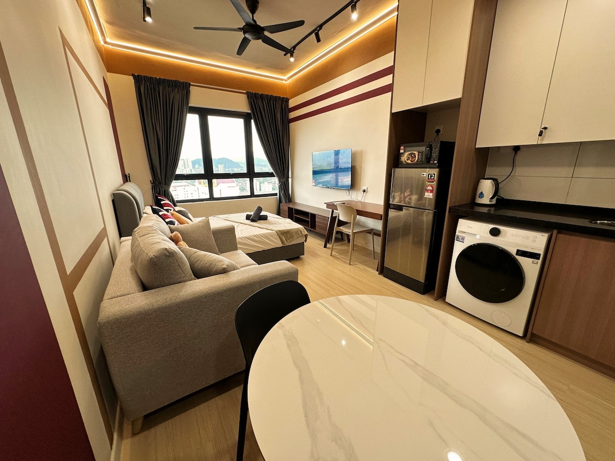 A modern studio interior exhibits a comfortable seating area with a light-colored sofa facing a wall-mounted TV. A round dining table is positioned nearby, and a kitchenette features a refrigerator and a washer-dryer unit. Large windows allow natural light to fill the space.