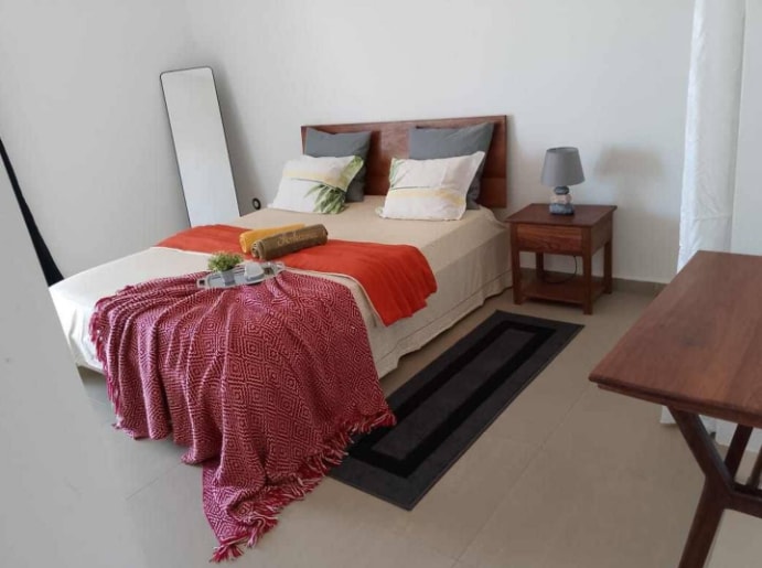A well-lit bedroom features a spacious bed adorned with neutral bedding and decorative pillows. A red throw blanket is draped over the foot of the bed. A small bedside table holds a lamp, while a decorative mirror leans against the wall, adding depth to the space.