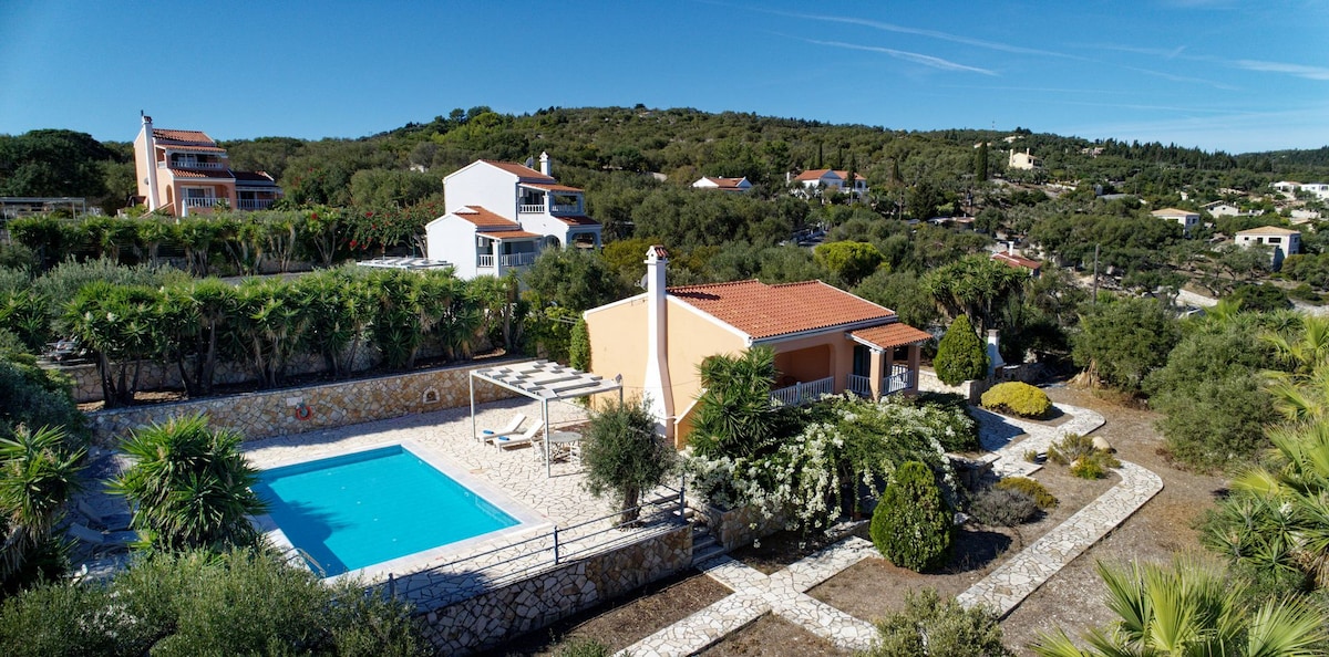 A spacious outdoor area is visible, featuring a rectangular pool surrounded by paved stone. Lush gardens with various green plants and trees are present, and a few lounge chairs are positioned at the poolside. The distant hills provide a serene backdrop to the property.