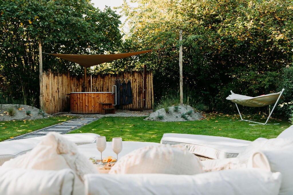 A serene garden space is highlighted by a wooden hottub, surrounded by natural greenery. A comfortable lounge area with soft cushions is positioned nearby, offering relaxation. A hammocking seat sways gently to the side, and a shaded area provides a cozy retreat.