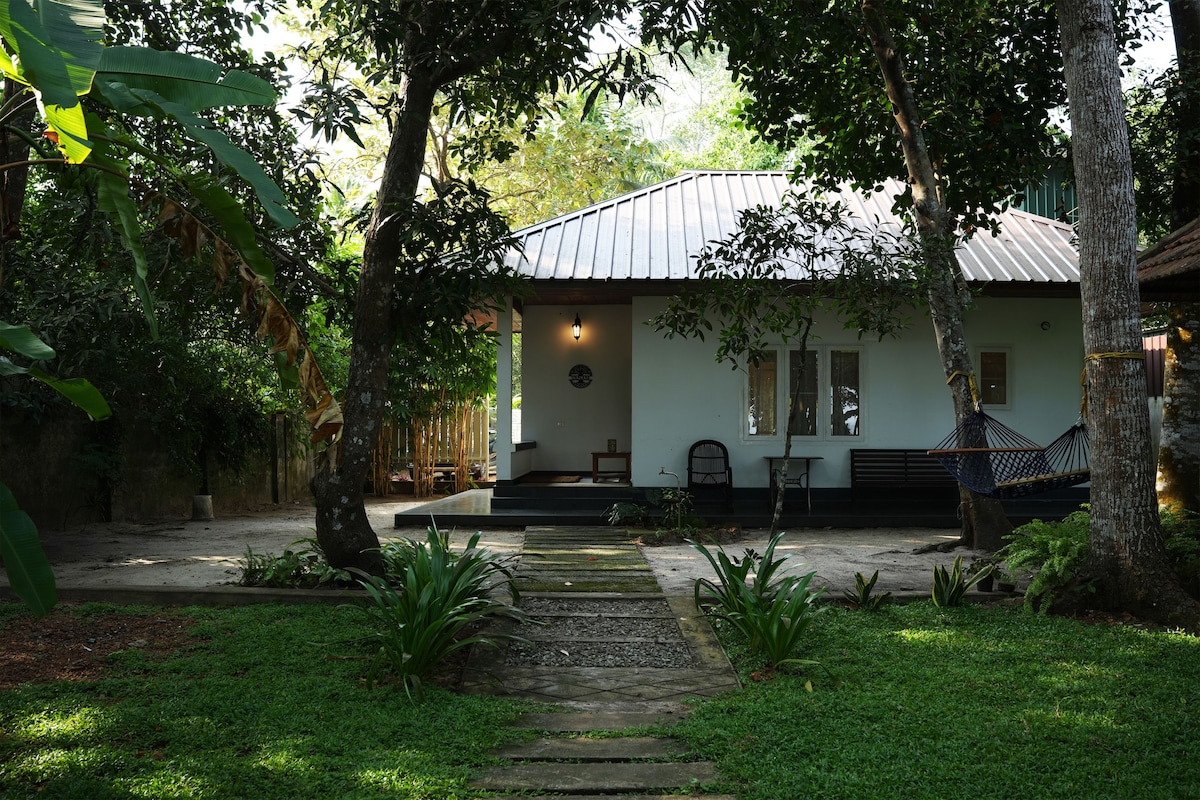 A serene villa is nestled among lush greenery, featuring a light-colored exterior with a sloped roof. A pathway lined with stones leads to the entrance, where seating arrangements are visible on a small porch surrounded by nature.