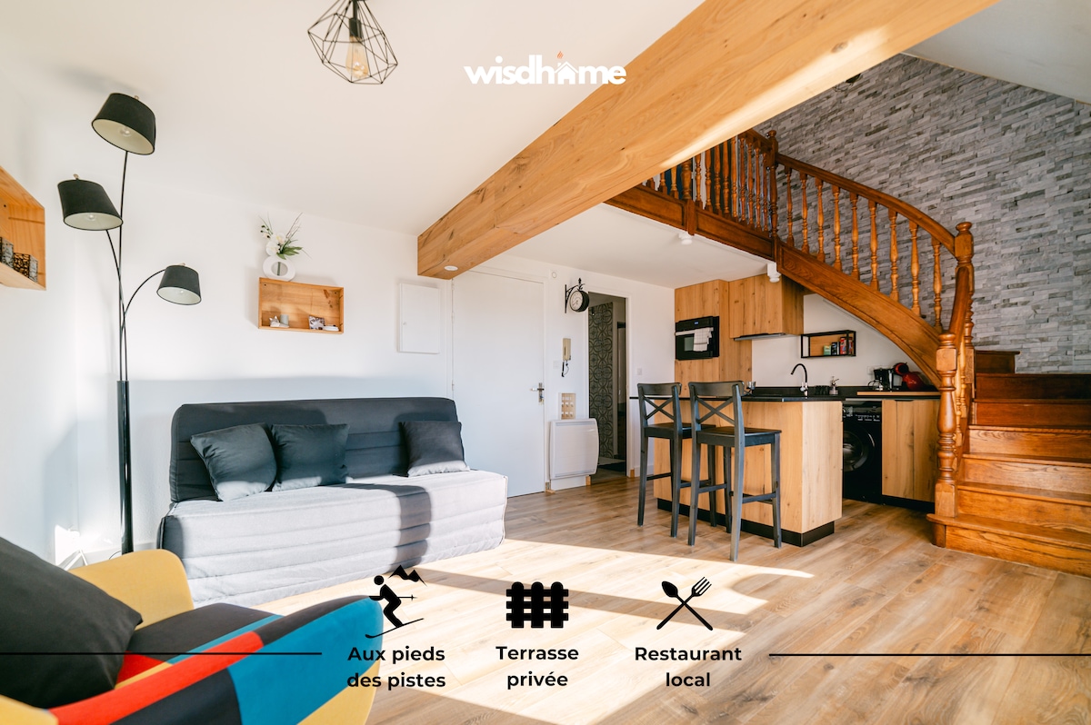 The living space features a convertible sofa, a dining area for four, and a wooden staircase leading to the upper level. A kitchenette is visible, equipped with essential appliances. Large windows allow natural light to flow into the room, enhancing the open atmosphere.