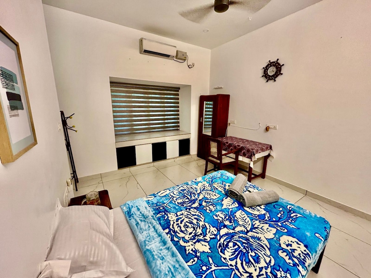 A spacious room is presented with a king-size bed adorned with a blue floral blanket. A window with layered blinds allows for soft light to filter in. A small working table and chair are positioned beside a storage unit, while a ceiling fan circulates the air.