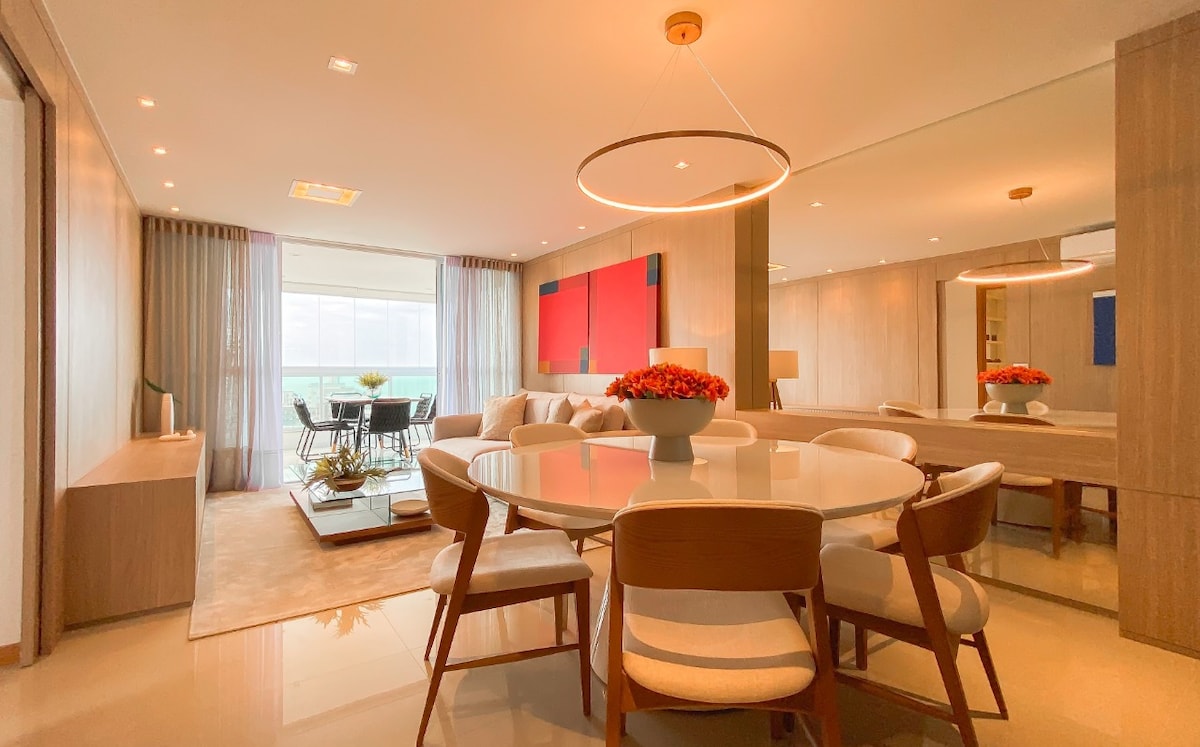 A contemporary dining area is shown, featuring a round table surrounded by six light-colored chairs. A stylish centerpiece of flowers is placed on the table. Large windows allow natural light to fill the space, while a comfortable seating area is visible in the background.