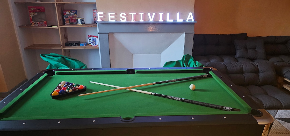 A game room features a green pool table with cues and balls positioned for play. Behind, a cozy seating area is complemented by the illuminated sign 'FESTIVILLA,' creating a welcoming ambiance. Shelves display various games, adding to the entertainment options.
