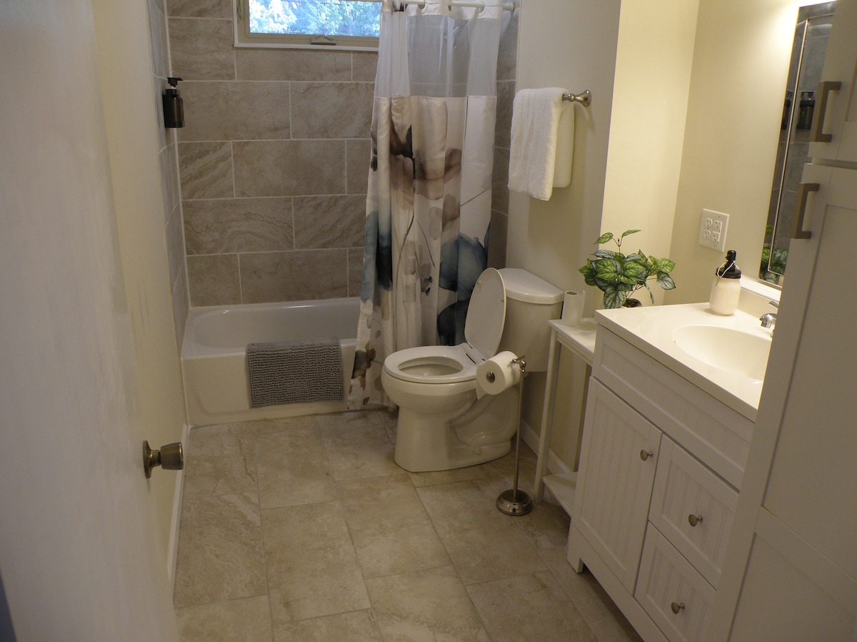 The bathroom features a clean design with light-colored tile flooring. A bathtub is located next to a shower with a floral curtain. A modern vanity with a sink is presented, accompanied by a mirror and decorative plant on the counter. Soft lighting enhances the space.