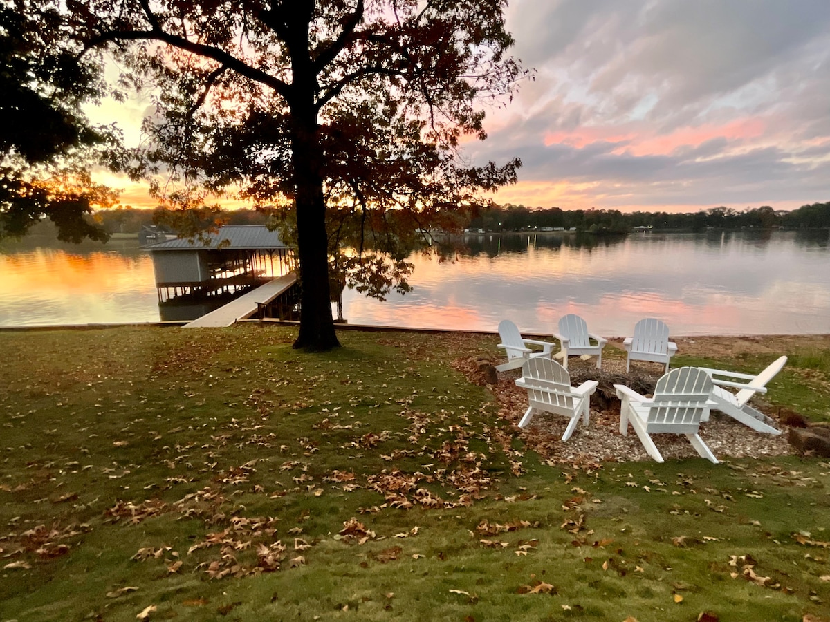 A serene lakeside scene features several white Adirondack chairs arranged in a circle on a grassy area. The calm water reflects a vibrant sunset, while a wooden boathouse and dock are visible in the distance, framed by trees with autumn foliage.