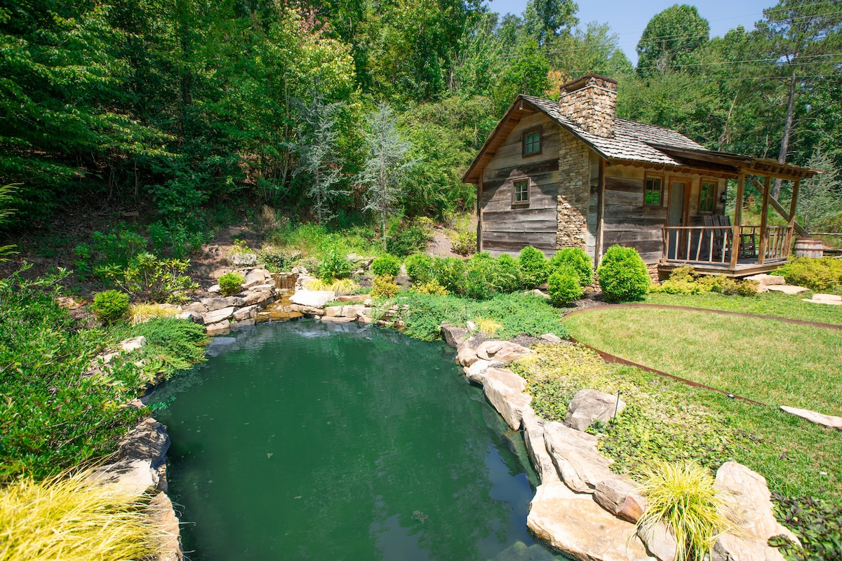 A rustic cabin is surrounded by lush greenery and a serene pond. Stone pathways lead to the cabin, which features a covered porch. Natural stones frame the pond, reflecting the lush landscape and bright blue sky.