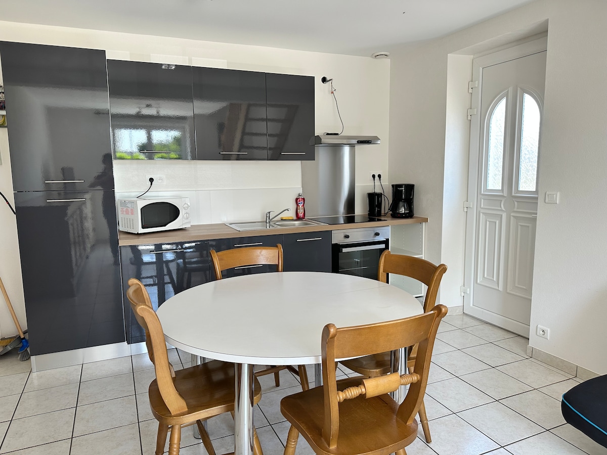 A modern kitchen area is displayed, featuring dark cabinetry and a white countertop. A round table with four wooden chairs is positioned centrally, while various appliances, including a microwave and coffee maker, are visible. Light enters through a nearby door, enhancing the space.
