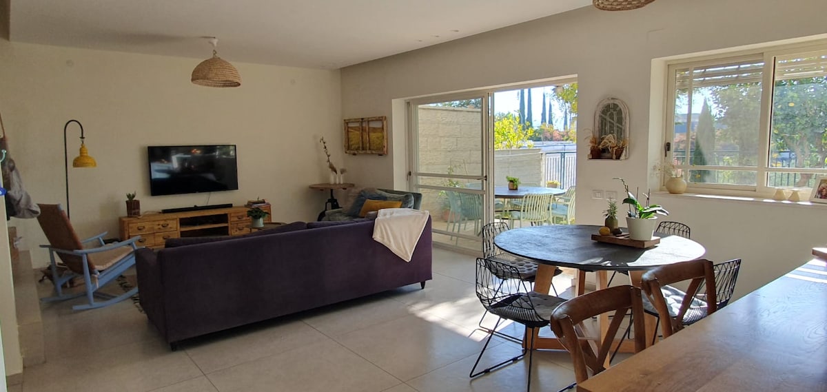 A spacious living area is illuminated by natural light from large glass doors. A comfortable sofa and a wooden coffee table are positioned near the TV. A dining table with wooden chairs is visible, featuring a centerpiece of plants. Outdoor seating can be seen beyond the glass doors.