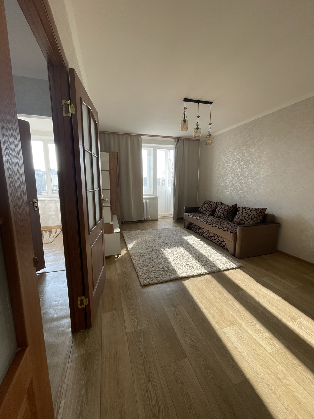 An inviting room features a light-colored wooden floor and a patterned sofa against the wall. Natural light floods in through a large window adorned with sheer curtains. A soft area rug is placed centrally, enhancing the room's warmth with minimal furnishings.